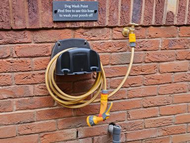 A coiled yellow garden hose attached to a faucet on a brick wall below a sign labeled Dog Wash Point