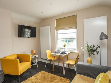 A living area with yellow armchairs a small dining table with yellow chairs a wall-mounted TV and a window with a brown blind at Cwm Gwyrfai in Caernarfon