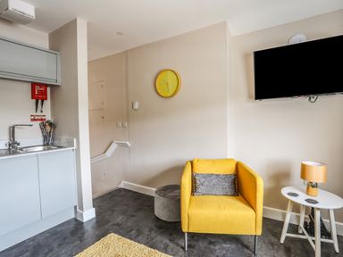 A living area with a yellow armchair a wall clock a small white table with a lamp and a kitchen sink at Cwm Gwyrfai Caernarfon