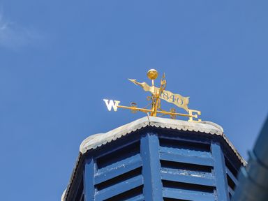 A weather vane with directions and the year 1840 on top of a blue wooden structure under a clear sky at Cwm Gwyrfai in Caernarfon