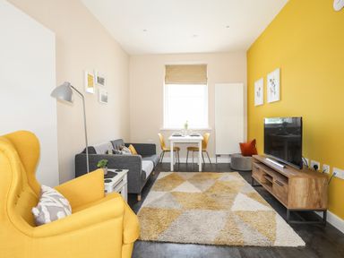A living room with a yellow armchair a gray sofa a small dining table with yellow chairs a patterned rug and a wooden TV stand at Moel Tryfan in Caernarfon