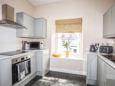 A kitchen with grey cabinets a microwave oven kettle toaster and a window with a vase of flowers and a cushion on the sill at Moel Tryfan Caernarfon