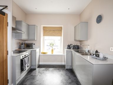 A kitchen with gray cabinets and countertops a window with a beige blind and a clock on the wall at Moel Tryfan in Caernarfon