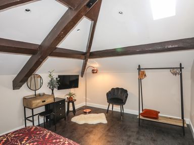 A bedroom with exposed wooden beams a vanity table with mirror a mounted TV a floor lamp a black chair and an open wardrobe with hangers at Moel Tryfan in Caernarfon