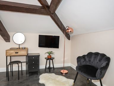 An attic room with a black chair a wooden vanity table with a round mirror a black chest of drawers a small round black table with a plant a copper floor lamp a wall mounted TV and a white rug on the floor at Moel Tryfan in Caernarfon