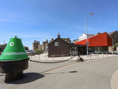 A green buoy with Caernarfon written on it near a small building and a red modern building with a castle in the background in Caernarfon