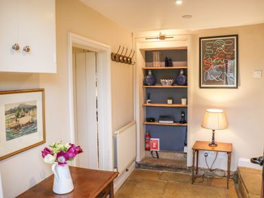 A hallway with wooden table and vase with flowers a wall-mounted coat rack shelves with decorative items a small table with a lamp and framed artworks at Manor Cottage in Poulton