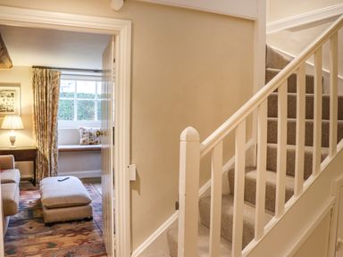A carpeted staircase with white railing next to a doorway leading to a living room with a sofa ottoman and a window at Manor Cottage in Poulton