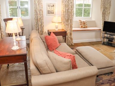 A living room with a beige sofa and cushions a wooden table with two lamps curtains windows and a television at Manor Cottage in Poulton