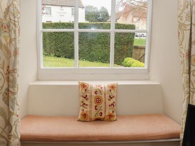 A window seat with an orange cushion and patterned pillow next to floral curtains overlooking a garden and a building with The Falcon Inn sign at Manor Cottage in Poulton