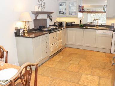 A kitchen with cream cabinets dark countertops a cream stove a sink and wooden chairs at Manor Cottage in Poulton