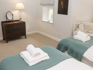A bedroom with two single beds with folded towels and green throws a wooden chest of drawers with a lamp and a mirror and a window with a painting on the wall at Manor Cottage in Poulton