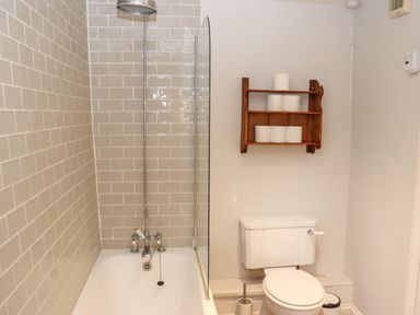 A bathroom with a bathtub shower combination and a toilet with toilet paper on a wooden shelf above at Manor Cottage in Poulton