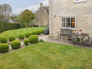 A garden with trimmed bushes a metal bench and table with chairs outside a stone house at Manor Cottage in Poulton