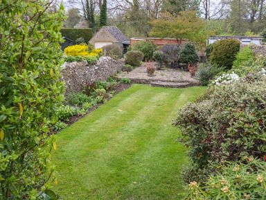 A garden with a central lawn bordered by bushes stone walls and outbuildings at Manor Cottage in Poulton