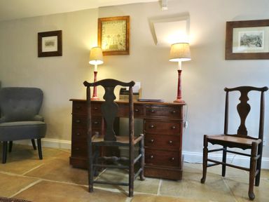 A room with a wooden desk with two red lamps two wooden chairs a grey armchair and framed pictures on the wall at Manor Cottage in Poulton