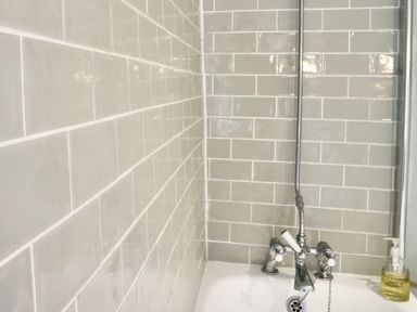 A bathroom corner with grey tiled walls a bathtub with chrome taps and a bottle of hand soap at Manor Cottage in Poulton