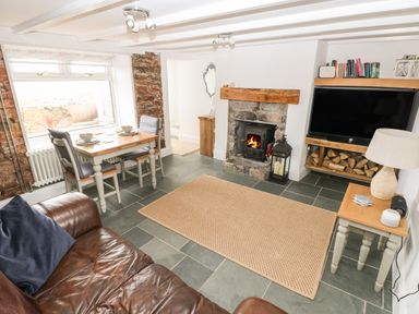 A living room with a brown leather sofa a dining table with two chairs a stone fireplace with a wood stove a television with firewood underneath and a small side table with a lamp at 1 Salt Cottage in Port Eynon