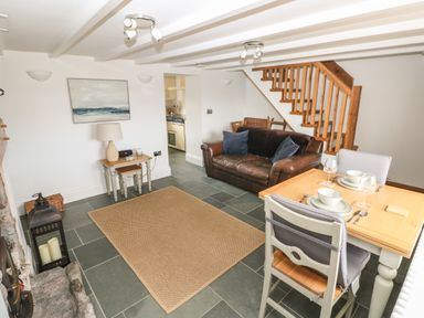 A living room with a brown leather sofa a wooden dining table with two place settings a small side table with a lamp and stairs in the background at 1 Salt Cottage in Port Eynon