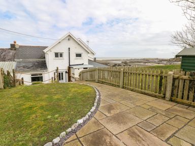 A backyard with a lawn and paved patio next to a white house with a view of the rocky shoreline at 1 Salt Cottage in Port Eynon