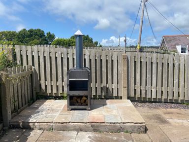 A wood-burning stove with firewood on a stone patio next to a wooden fence at 1 Salt Cottage in Port Eynon