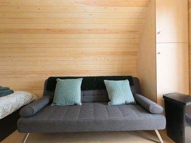 A gray sofa with two blue cushions against a wood-paneled wall with a bed and cabinet nearby at Green Fields in St Buryan