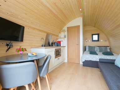 An interior room with a round dining table and chairs a kitchenette with a microwave and utensils a bed with pillows and towels and a sofa at Green Fields in St Buryan