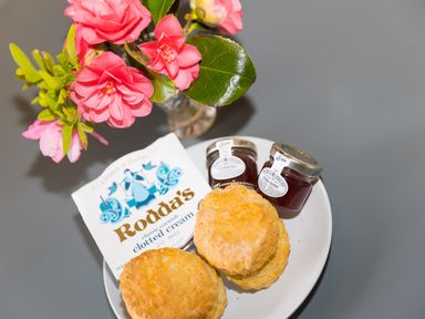 A plate with two scones two jars of strawberry jam and a pack of clotted cream next to a vase with pink flowers at Green Fields in St Buryan