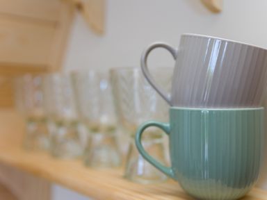 Two ceramic mugs stacked on a wooden shelf with glass cups in the background at Green Fields in St Buryan