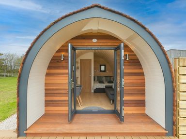 A wooden pod with open glass doors showing a bed and seating area inside at Green Fields in St Buryan