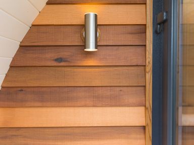 A wooden paneled wall with a modern silver cylindrical light fixture mounted above a dark framed window at Green Fields in St Buryan