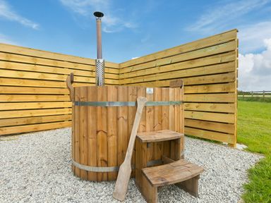 An outdoor wooden hot tub with a stove pipe and wooden steps on gravel surrounded by wooden fencing at Green Fields in St Buryan