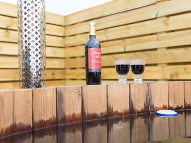 A bottle of red wine and two glasses filled with wine on the edge of a wooden hot tub in an outdoor fenced area at Green Fields in St Buryan