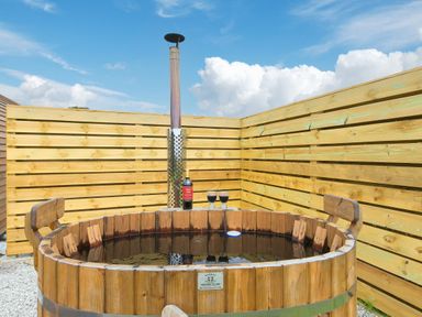 An outdoor wooden hot tub with two seats and a wine bottle with glasses on the edge at Green Fields in St Buryan