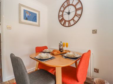 A dining area with a wooden table set for four with bowls and plates orange and gray chairs and wall clock at Robellen Corner in Adderbury