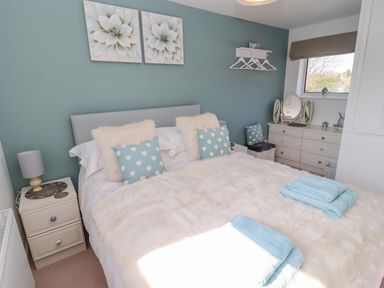 A bedroom with a bed with pillows and a fur blanket two bedside tables a dresser with a mirror and a window at Robellen Corner in Adderbury