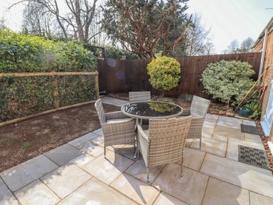 A patio with a round glass table and four wicker chairs surrounded by plants and a wooden fence at Robellen Corner in Adderbury