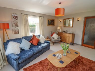 A living room with a blue sofa and an armchair with cushions a wooden coffee table with flowers and coasters and a sideboard with decorations at Robellen Corner in Adderbury