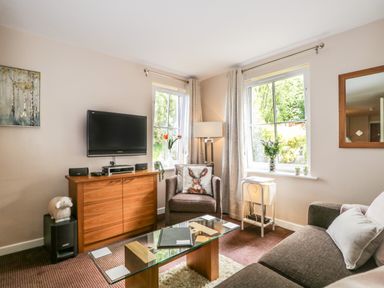 A living room with a brown sofa, armchair, glass coffee table, wooden cabinet with TV and two windows at Cosy Corner in the Lakes in Greystoke