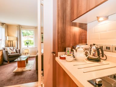 A kitchen counter with a kettle, teapot, toaster, and cups next to a living room with armchairs and a coffee table at Cosy Corner in the Lakes in Greystoke