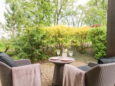 An outdoor patio area with two wicker chairs a small table with plates a jug and glasses surrounded by green bushes at Cosy Corner in the Lakes in Greystoke