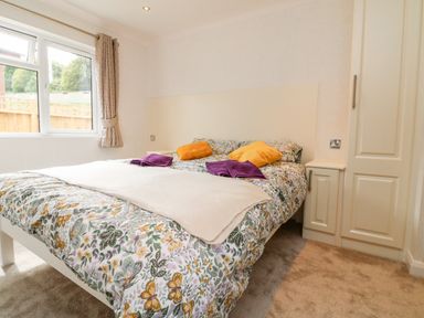 A bedroom with a bed covered with floral bedding two orange pillows and two purple towels near a window with beige curtains and built-in wardrobes at Ridge's Retreat No 7 Kigbeare Lodges Ashbury Resort near Fowley Cross Sourton Down and Okehampton