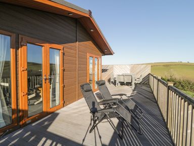 An outdoor wooden deck with two black lounge chairs and a table with chairs overlooking grassy hills at Ridges Retreat No 7 Kigbeare Lodges Ashbury Resort near Fowley Cross Sourton Down and Okehampton