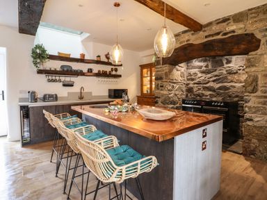 A kitchen with a copper countertop island with wicker stools and stone wall with a stove at Plas Glan Yr Afon in Llangernyw
