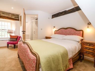 A bedroom with a bed with floral upholstery and a green blanket a wooden nightstand a pink chair near a window and an open door showing a tiled shower at Plas Glan Yr Afon in Llangernyw