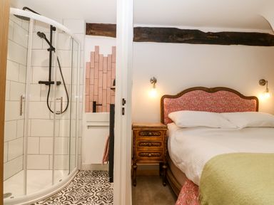 A bedroom with a floral headboard bed and bedside table next to a bathroom with a shower and patterned floor tiles at Plas Glan Yr Afon in Llangernyw