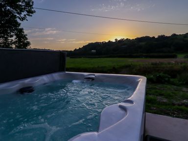 An outdoor hot tub with bubbling water overlooking a field and hills at sunset at Plas Glan Yr Afon in Llangernyw