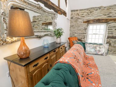 A living area with a green sofa covered with a patterned orange blanket next to a wooden sideboard with a lamp plant and glass vase on top and a large ornate mirror on the wall at Plas Glan Yr Afon in Llangernyw