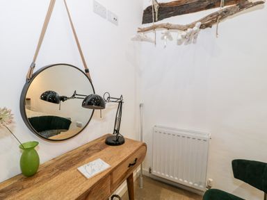 A wooden desk with a black desk lamp green vase and notebook below a round hanging mirror in a corner with a radiator and green chair at Plas Glan Yr Afon in Llangernyw