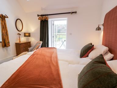 A bedroom with two single beds covered in white bedding and orange throw a wooden dresser with a lamp and chair near a door leading outside at Plas Glan Yr Afon in Llangernyw
